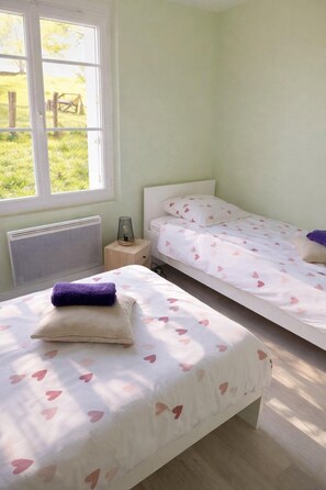 A bedroom with two beds, a radiator, a bedside table with a lamp, and a window with a view of the outdoors. - The source - Le Grand-Pressigny (Le Grand-Pressigny)