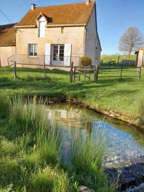 A house with a small stream in front, surrounded by grass and a wooden fence. - The source - Le Grand-Pressigny (Le Grand-Pressigny)