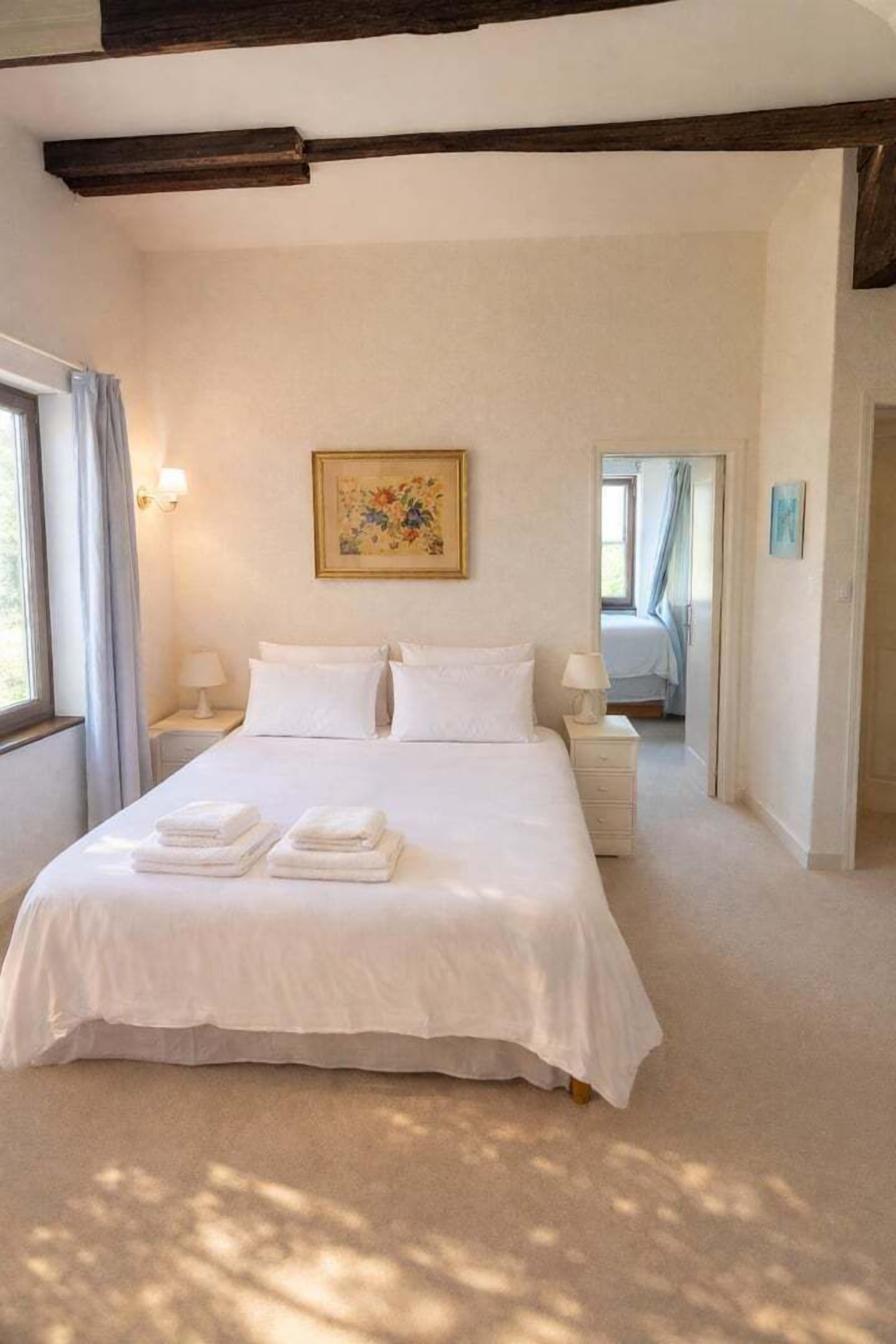 A bedroom with a large bed, two bedside tables, a painting on the wall, and a window with curtains.
