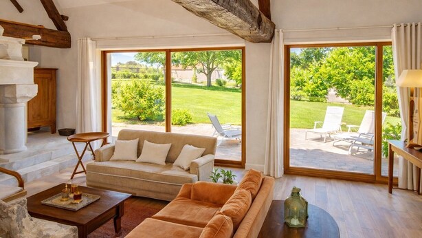 Ein geräumiges Wohnzimmer mit einem beigen Sofa, einem Holztisch und einem großen Fenster mit Blick auf einen Garten und Sitzgelegenheiten im Freien.