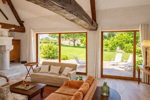 Ein geräumiges Wohnzimmer mit einem beigen Sofa, einem Holztisch und einem großen Fenster mit Blick auf einen Garten und Sitzgelegenheiten im Freien.