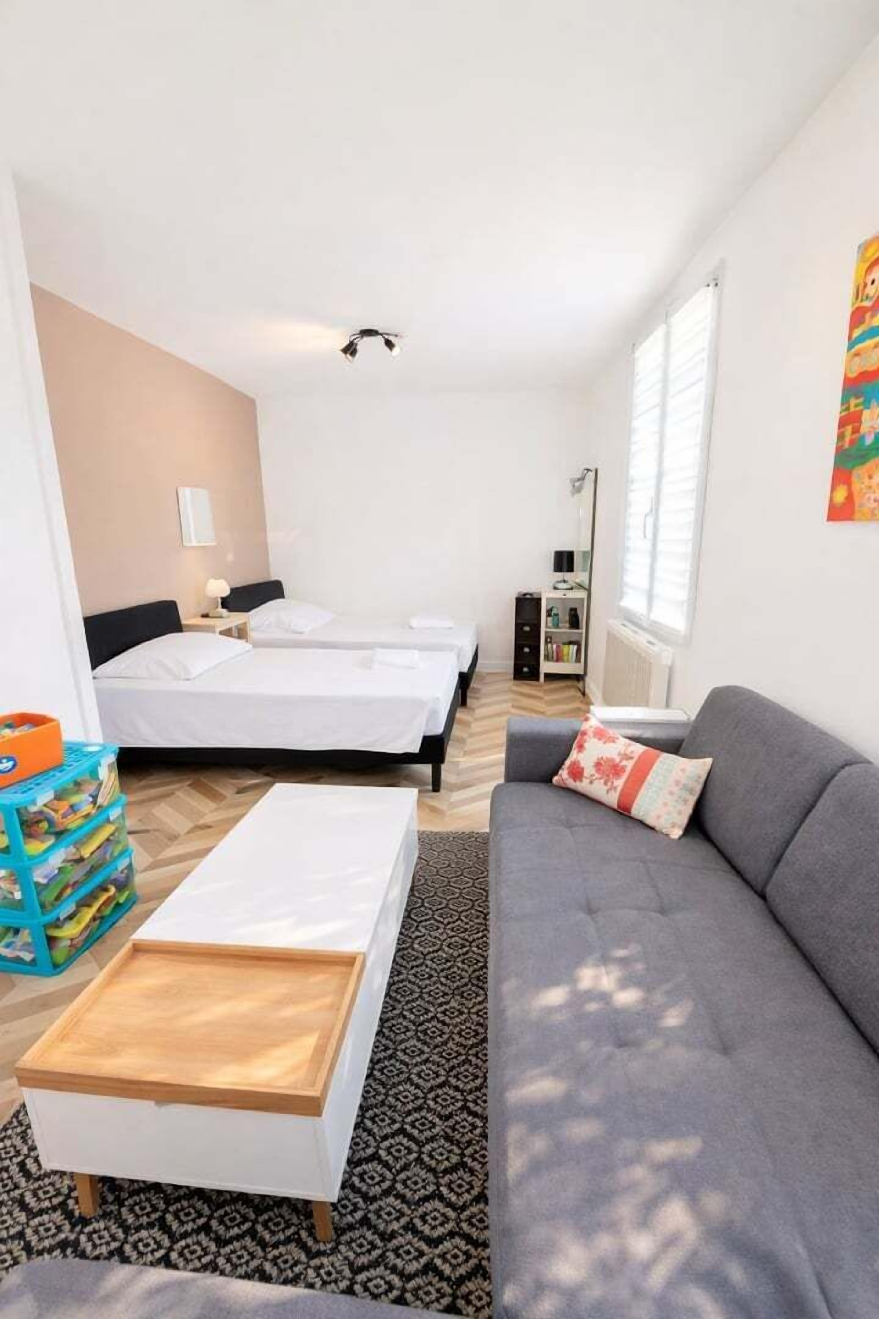 A room with two beds, a grey sofa, a wooden side table, a patterned rug, and a small bookshelf.