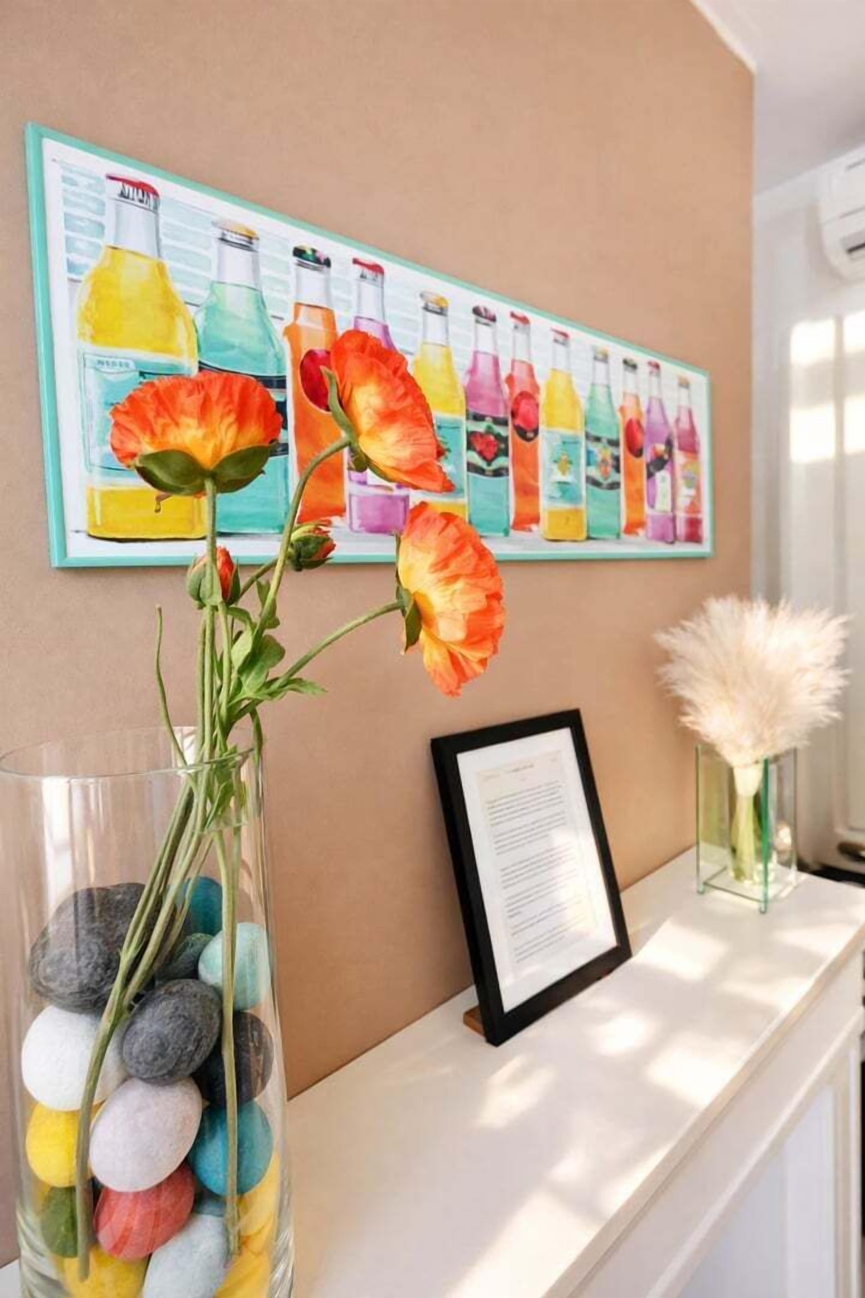 Una zona de recepción con una obra de arte enmarcada que muestra botellas de colores, un jarrón con flores naranjas, un documento enmarcado y un jarrón de vidrio con plumas blancas.