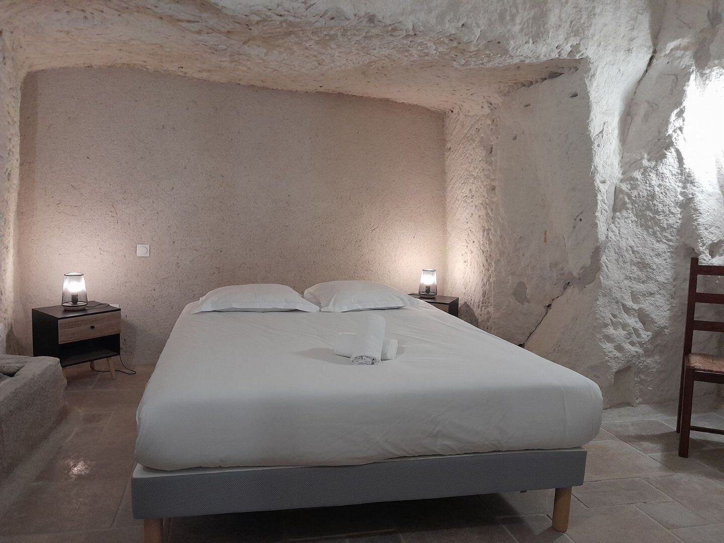 Un dormitorio con una cama, dos mesitas de noche con lámparas, una silla de madera y una pared de piedra.