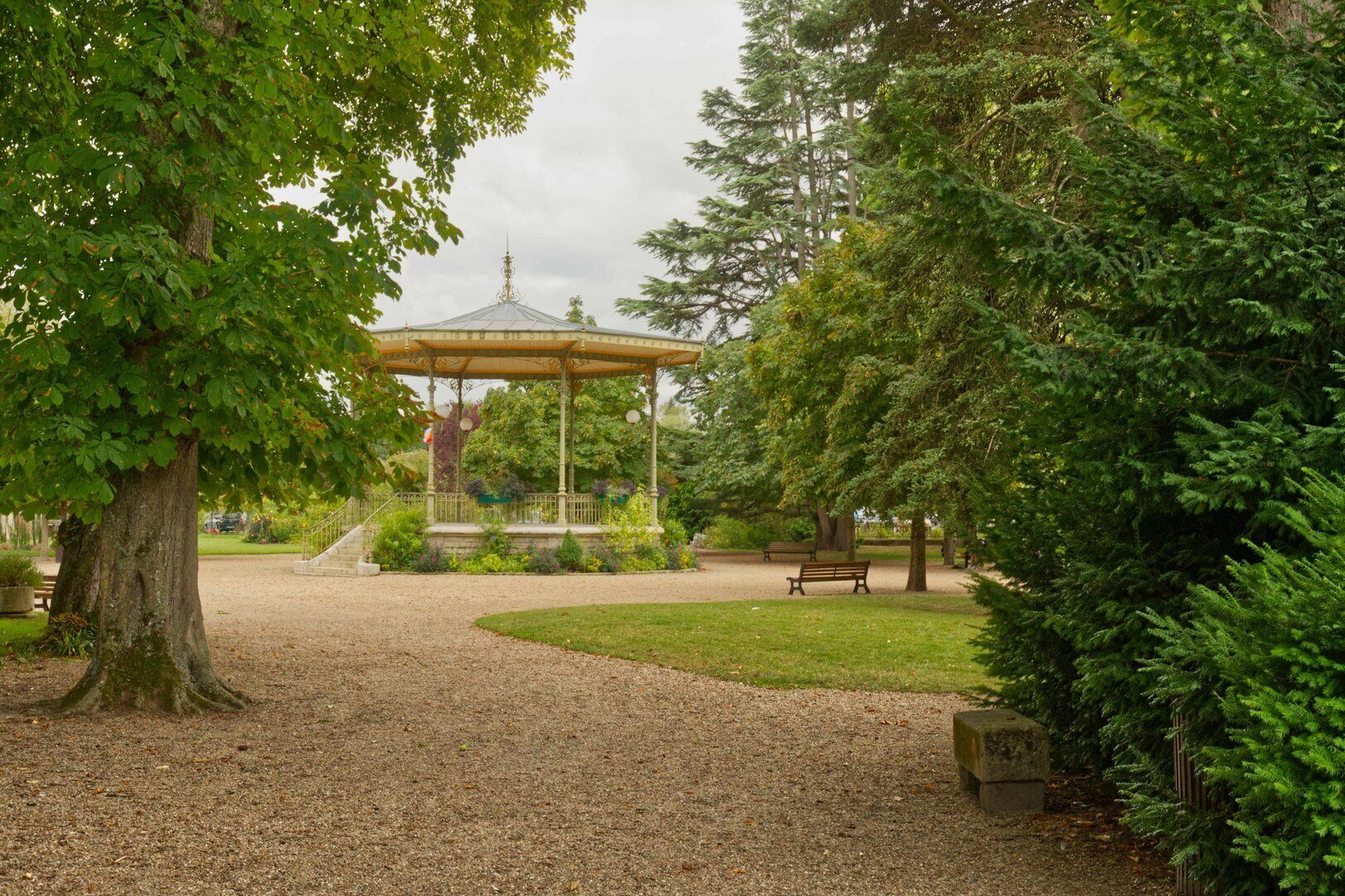 Een prieeltje in een park met een grindpad, bankjes en weelderige begroeiing.