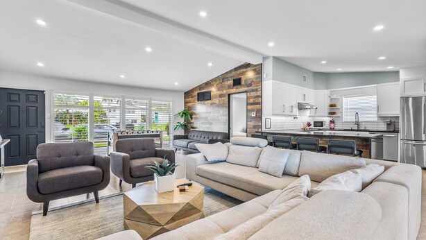 A modern living room with a large sectional sofa, a coffee table, and two armchairs. A kitchen with white cabinets and stainless steel appliances is visible in the background. - Waterfront Ft Lauderdale Retreat | Kayaks Included (Fort Lauderdale)
