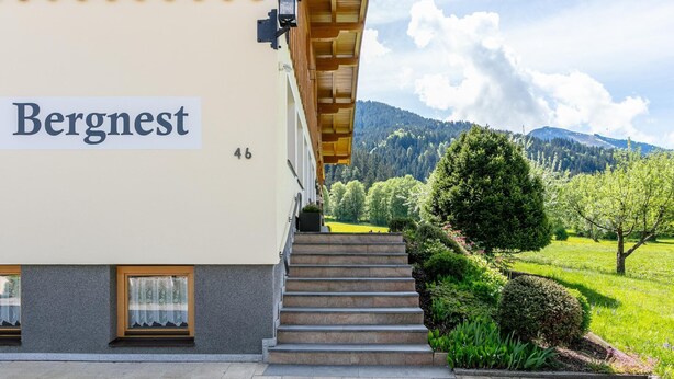 Un edificio llamado 'Bergnest' con una escalera que conduce a un vestíbulo cubierto, rodeado de vegetación y un paisaje montañoso.