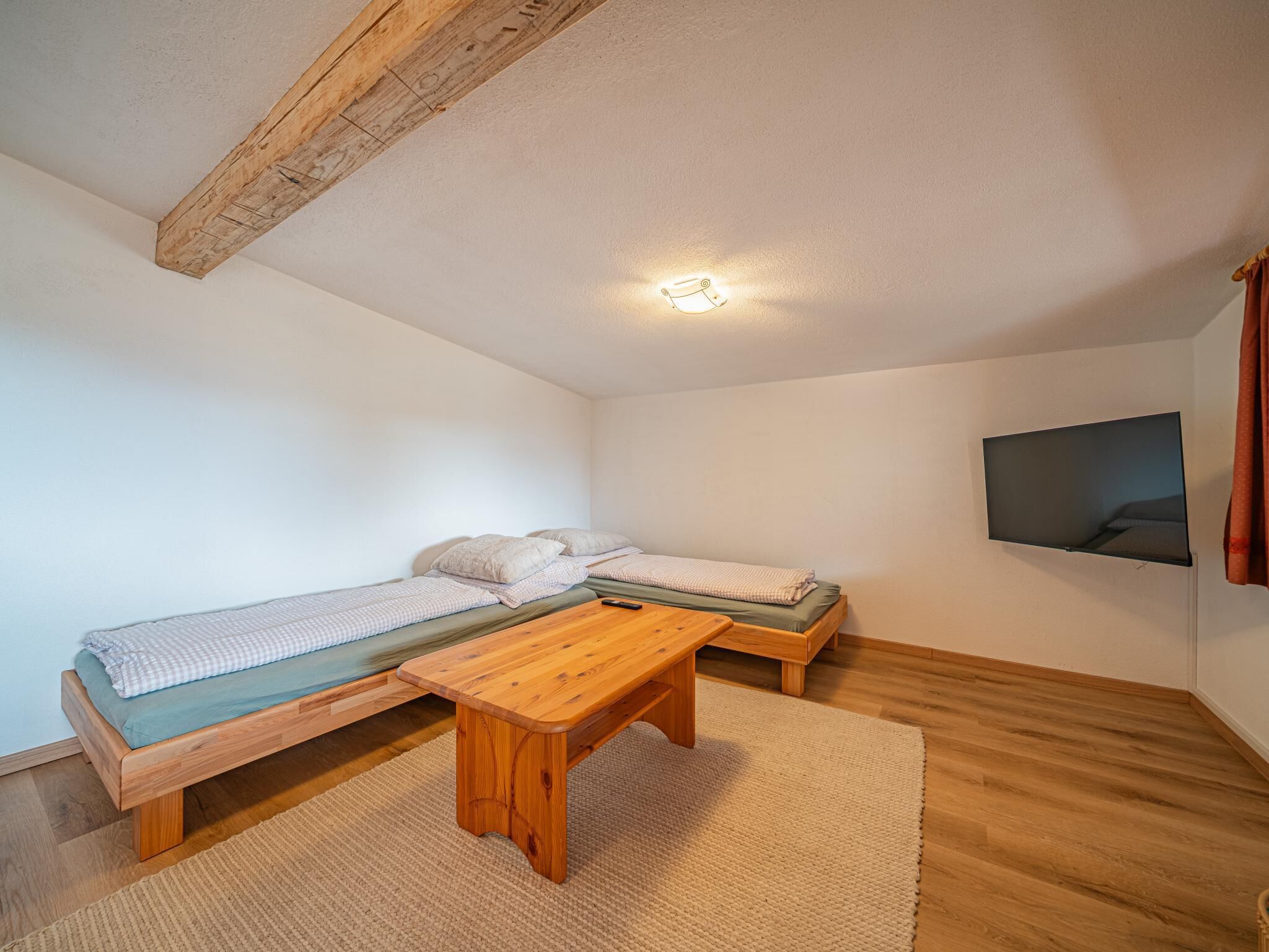 A small, cozy room with a wooden ceiling, a bed with a green mattress, a bench, a small table, and a TV mounted on the wall.