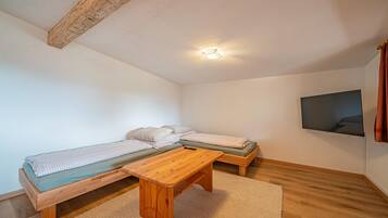 A small, cozy room with a wooden ceiling, a bed with a green mattress, a bench, a small table, and a TV mounted on the wall.