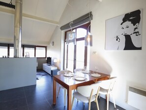 Un coin repas avec une table en bois dressée pour quatre personnes, une cuisine moderne en arrière-plan et une grande fenêtre laissant entrer la lumière naturelle.