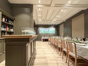 A long dining table with chairs, a bar area with bottles, and a large screen displaying an image.