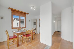 Un coin repas avec une table et des chaises en bois, une fenêtre avec des stores et une cuisine en arrière-plan.