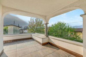 Un balcon avec un sol carrelé, une colonne en pierre et une vue sur des maisons et des arbres.