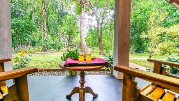 Une table en bois, recouverte d’une nappe rouge et agrémentée de tasses jaunes, est placée sur un balcon d’où l’on peut admirer un jardin luxuriant.