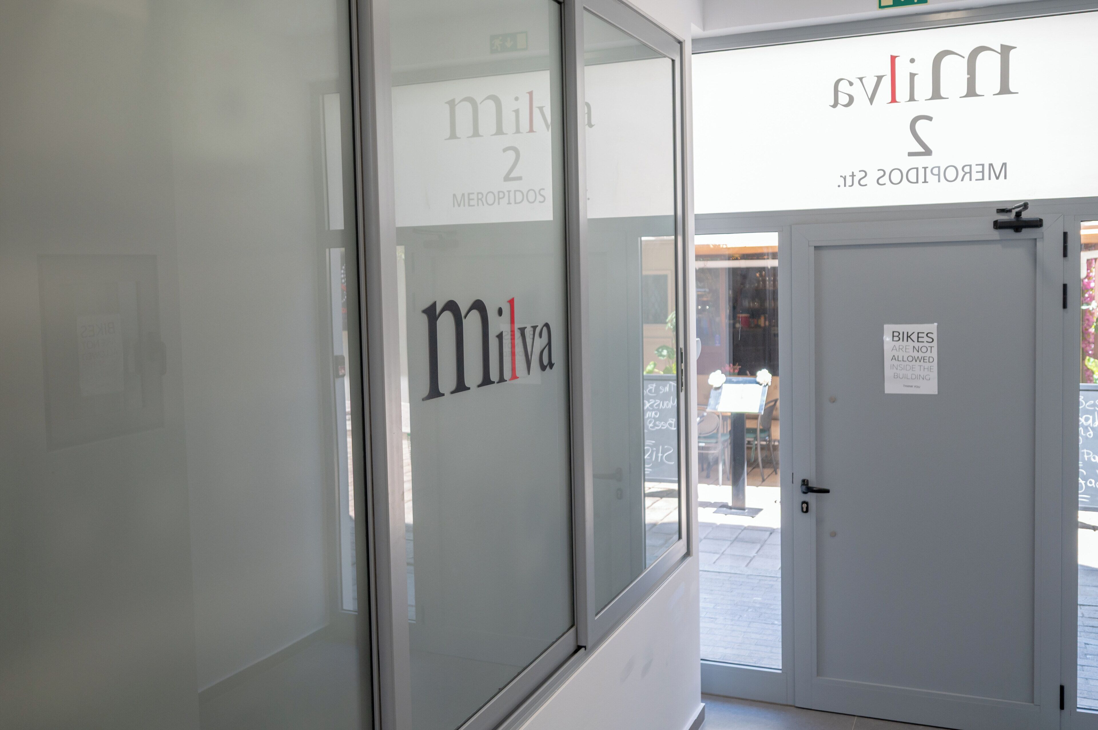 A glass door with the text 'Milva' and 'Meropidos St.' Another door has a sign stating 'Bikes are not allowed inside the building.'
