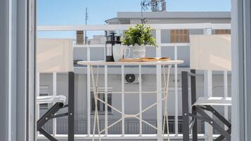 Un balcón con una mesa metálica blanca, una planta en maceta, un libro y una cafetera.