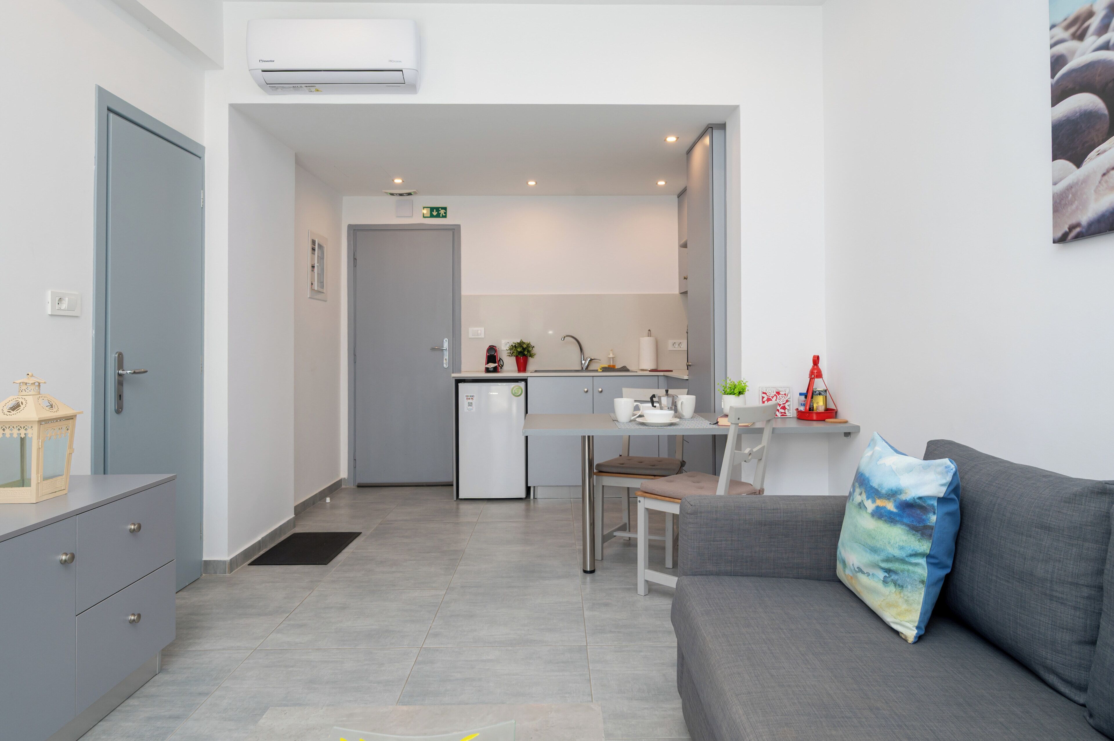 A modern kitchenette with a refrigerator, sink, and countertop. There is a dining area with a table and chairs, and a gray sofa with a decorative pillow.
