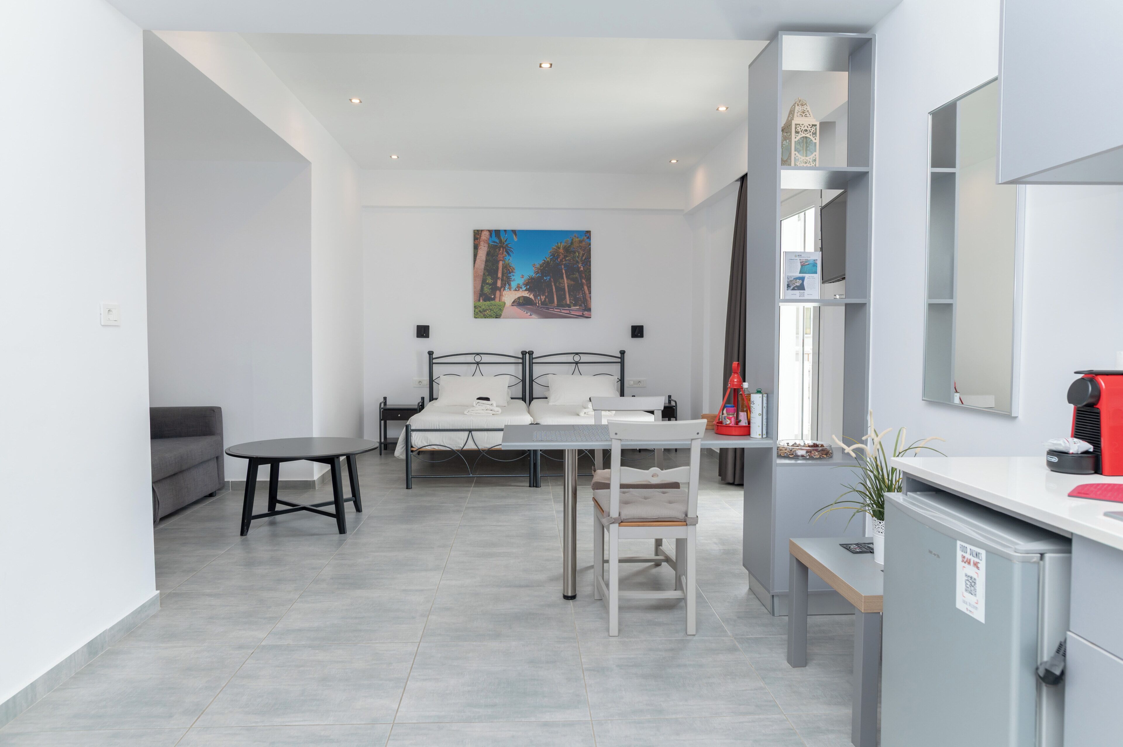 A modern hotel room with a bed, a desk, a chair, a small table, and a kitchenette area.