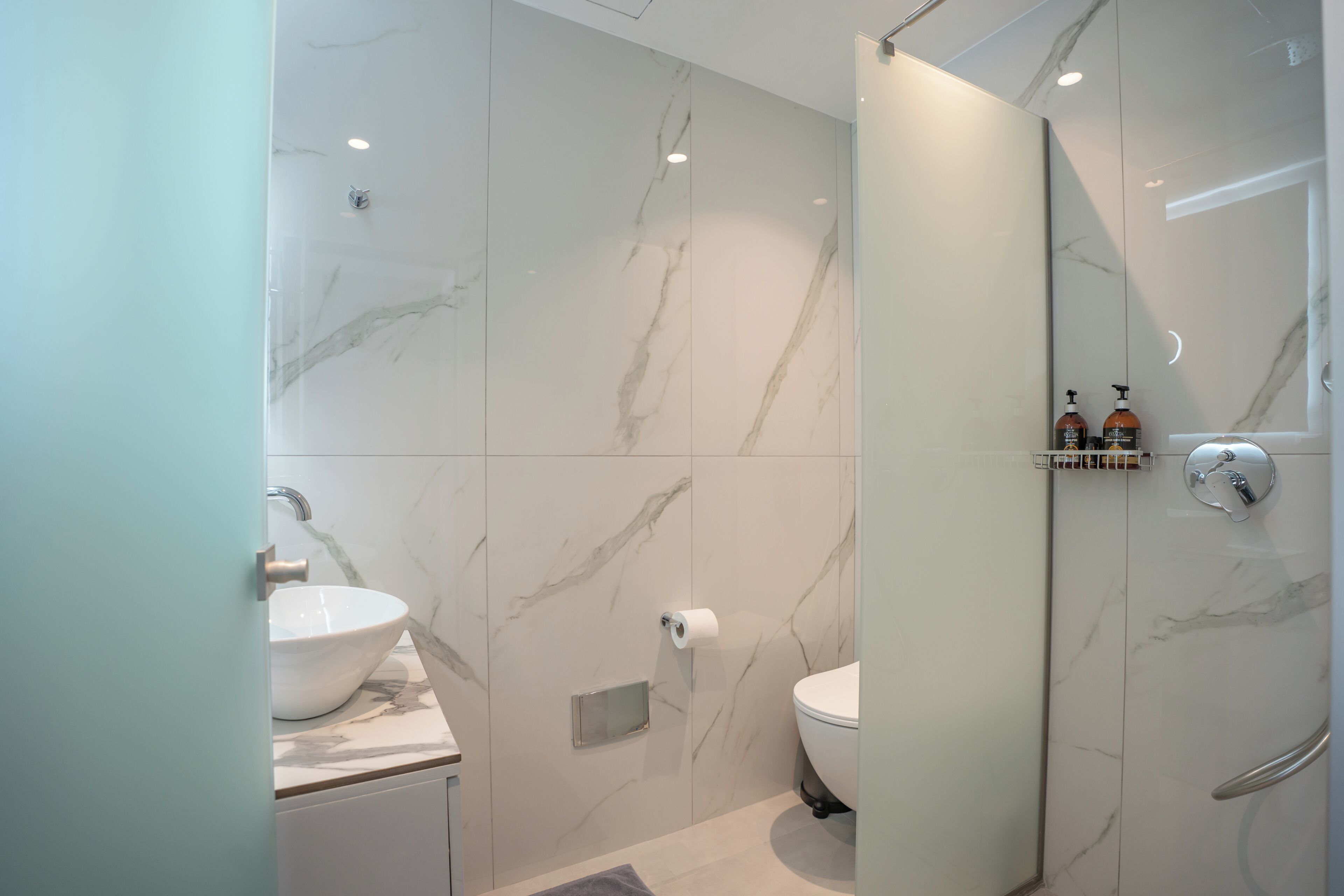 A modern bathroom with a marble wall, a white sink, a toilet, and a glass shower enclosure.