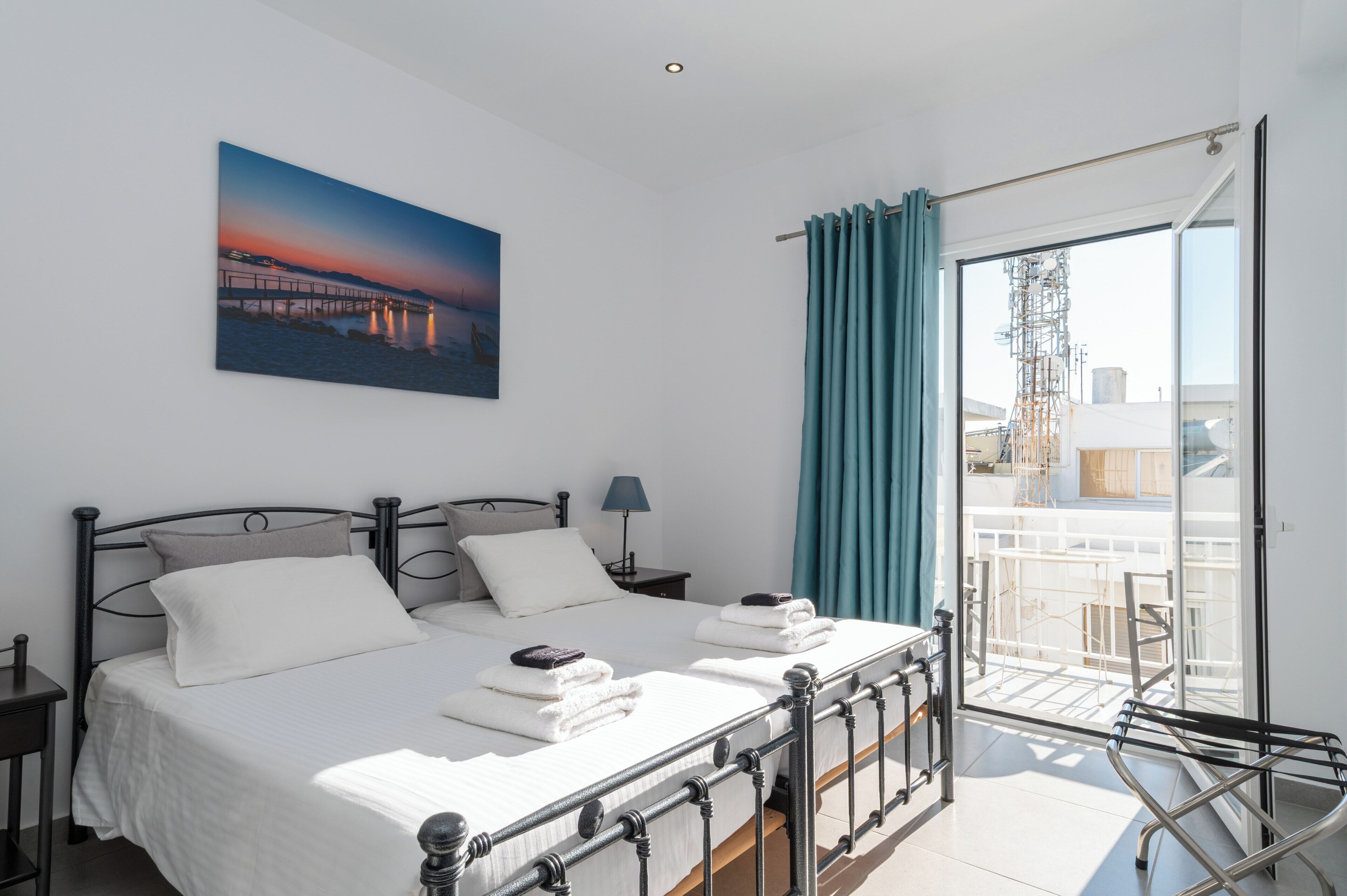 A neatly made bed with white linens, a nightstand with a lamp, a painting of a sunset on the wall, and a balcony with a view of the city.