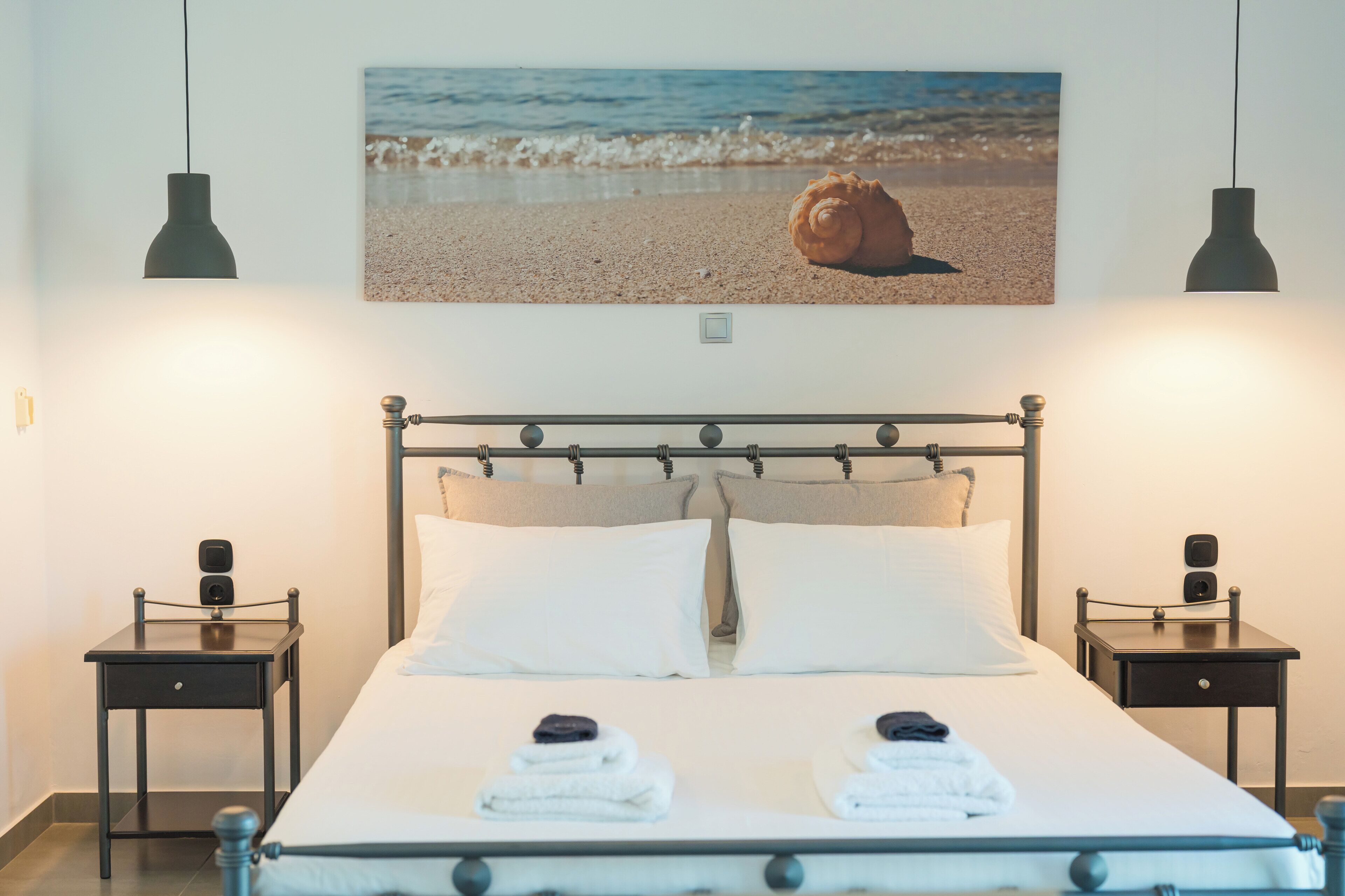 Un lit bien fait, avec deux oreillers, une table de chevet sur laquelle se trouve une lampe, et un tableau représentant une scène de plage, accroché au mur.