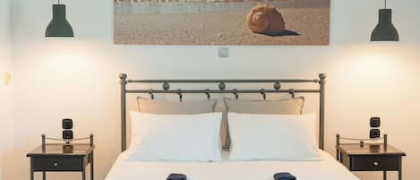 Un lit bien fait, avec deux oreillers, une table de chevet sur laquelle se trouve une lampe, et un tableau représentant une scène de plage, accroché au mur.