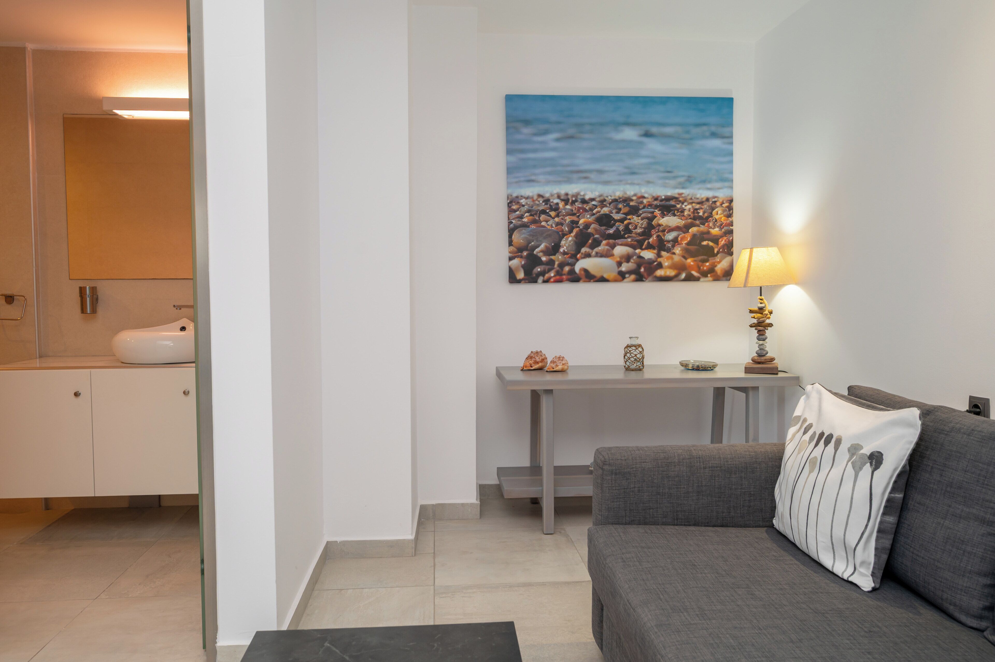 Una sala de estar moderna con un sofá gris, una mesita con una lámpara y un cuadro con una escena de playa en la pared.
