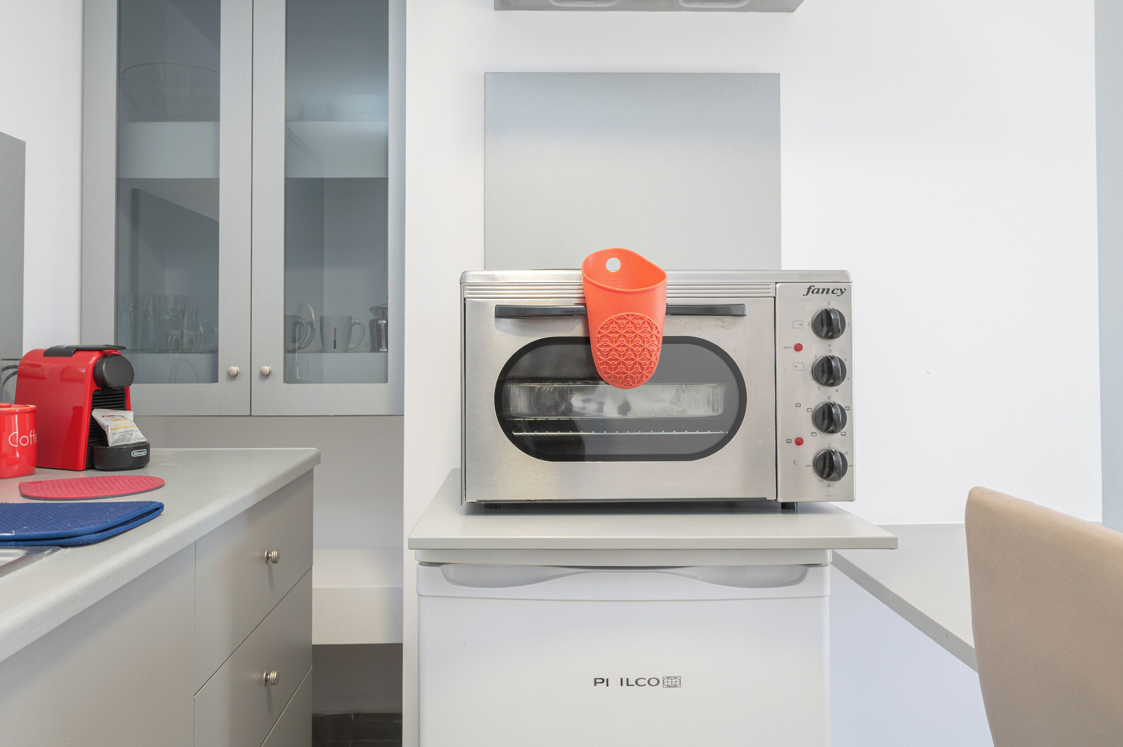 Una kitchenette con un horno tostador moderno, una cafetera roja y una tabla de cortar azul sobre el mostrador.