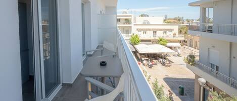 A balcony with a table and chairs, a view of a courtyard with outdoor seating, and a building with multiple floors and balconies.