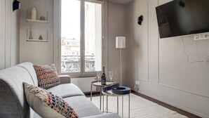 Un salon moderne avec un canapé, un téléviseur, une fenêtre donnant sur des immeubles et une petite table sur laquelle se trouvent une bouteille et un verre.