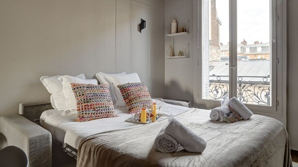 Une chambre d’hôtel comprenant un lit, des oreillers, une table de chevet avec des verres et une bouteille, une fenêtre donnant sur des immeubles et une étagère avec des objets décoratifs.