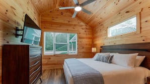 Une chambre dans une cabane en bois, comprenant un lit, une commode, un ventilateur de plafond et une fenêtre avec des stores.