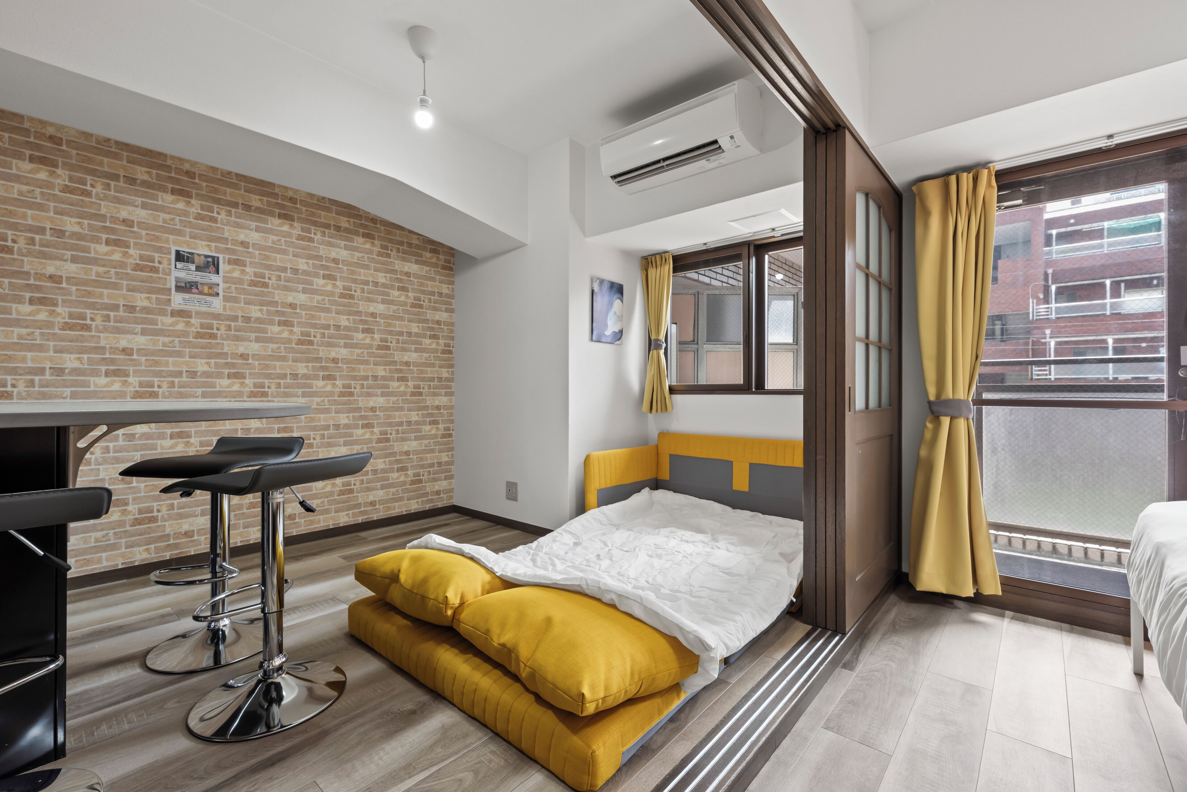 A hotel room with a bed, a small table with a chair, a window with curtains, and a brick wall.