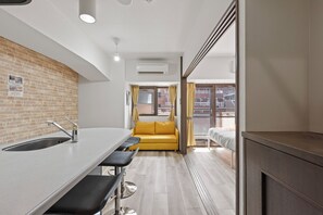 A modern kitchen with a sink, a dining area with a bar stool, a yellow sofa, and a view of the city through a window. - Grace Nagahori 402 (Osaka)