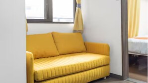 A small room with a yellow sofa, a window with curtains, and a bed with a white mattress. - Grace Nagahori 402 (Osaka)