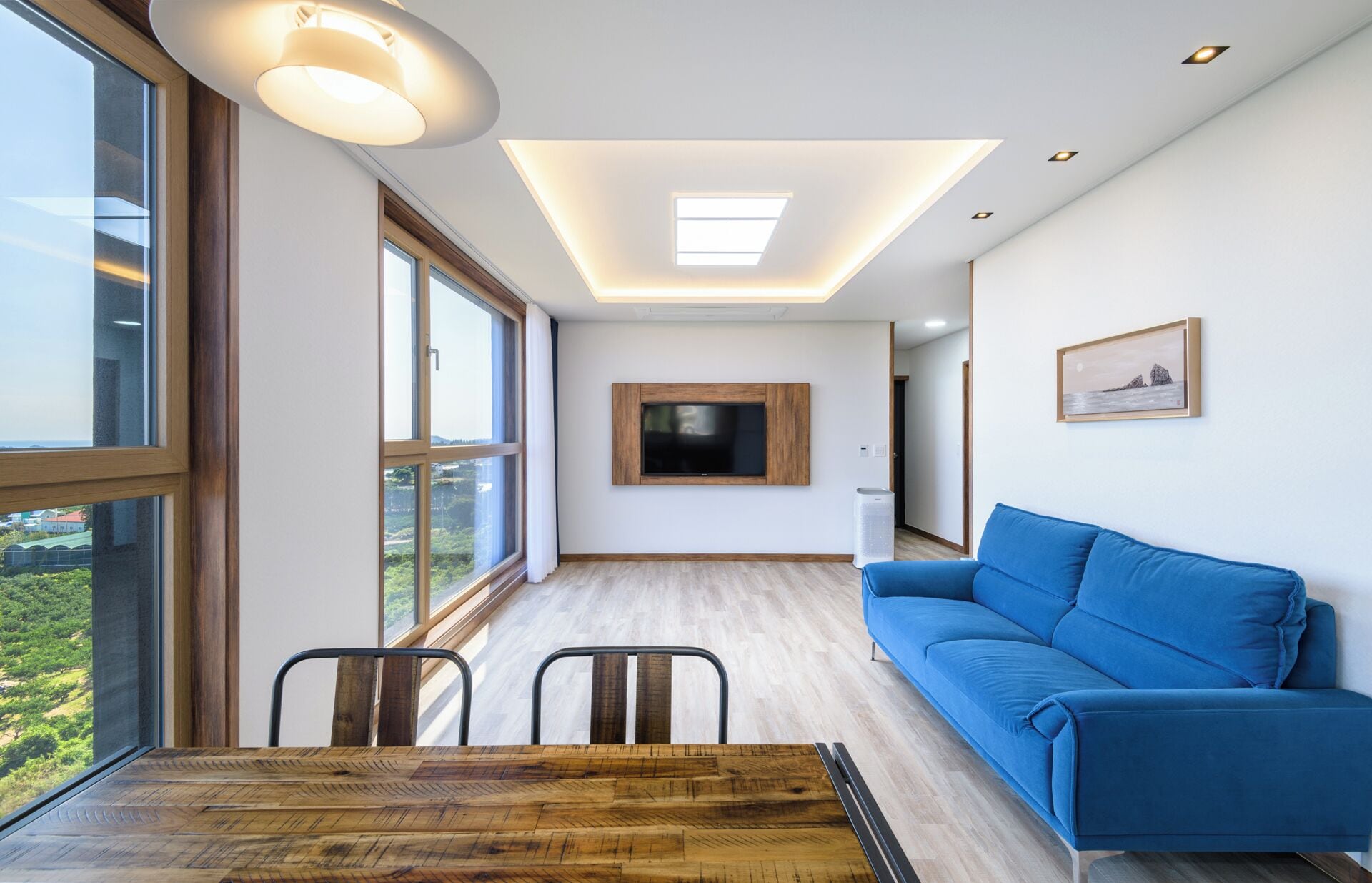 A modern living room with a large window, a blue sofa, a wooden dining table, and a flat-screen TV mounted on the wall.