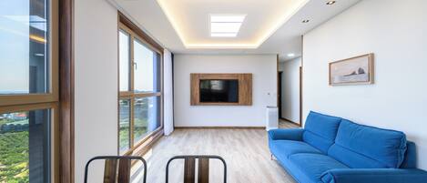 A modern living room with a large window, a blue sofa, a wooden dining table, and a flat-screen TV mounted on the wall.