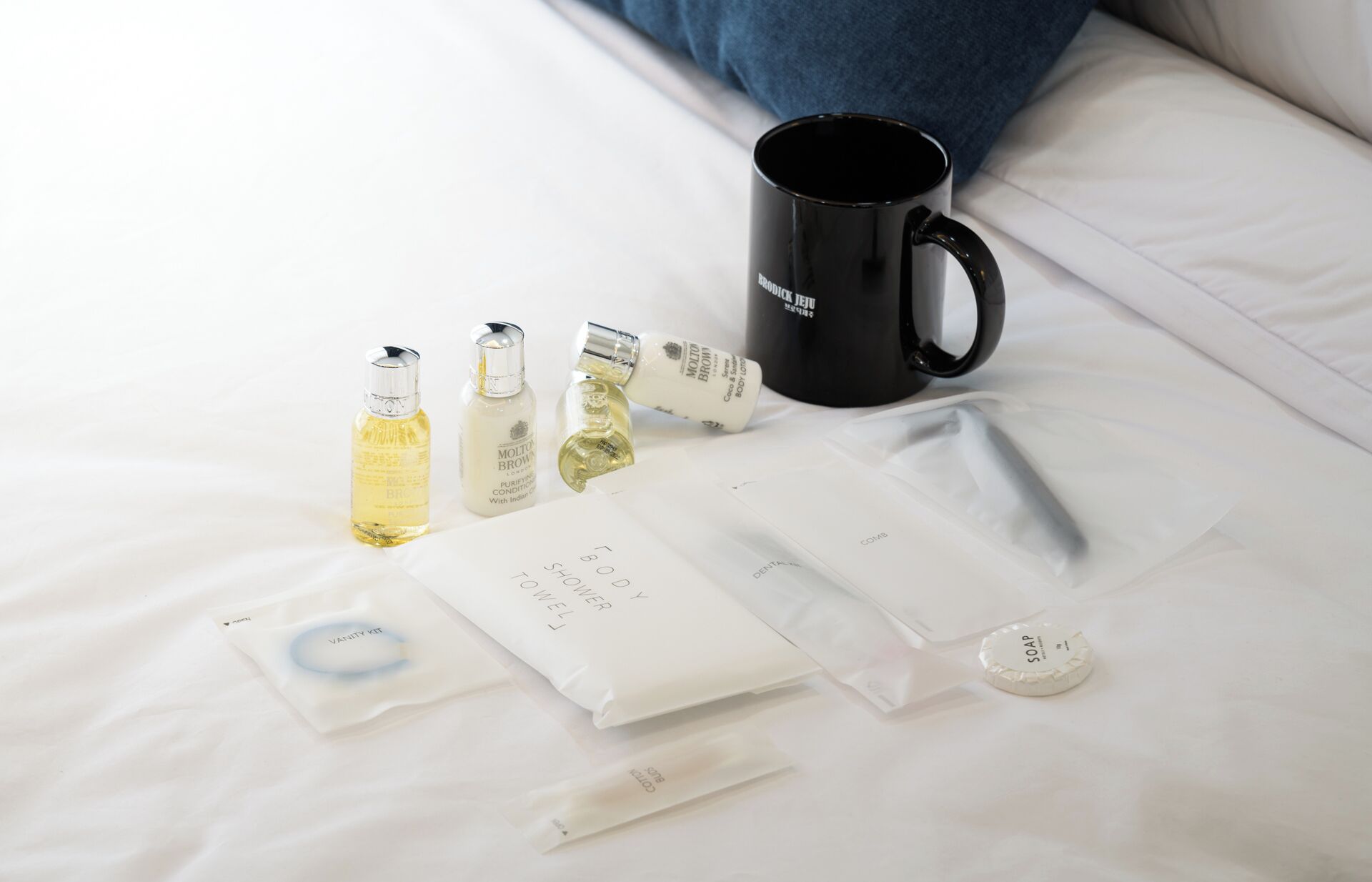 Toiletries on a bed, including a black mug, bottles of lotion, and a vanity kit.