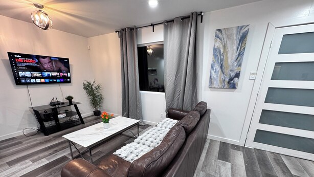 A modern living room with a leather sofa, a marble coffee table, a TV mounted on the wall, and a large window with curtains. - Compact 3 bed/1bath house 4 miles away from Downtown (Dallas)