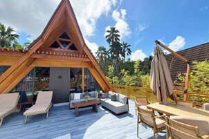 Un área de estar exterior moderna con una terraza de madera, una estructura triangular de madera y vistas al verde circundante.