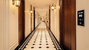 Un corridoio di un albergo con pavimento a scacchi bianchi e neri, lampade a parete e una porta con la targa numero 302.
