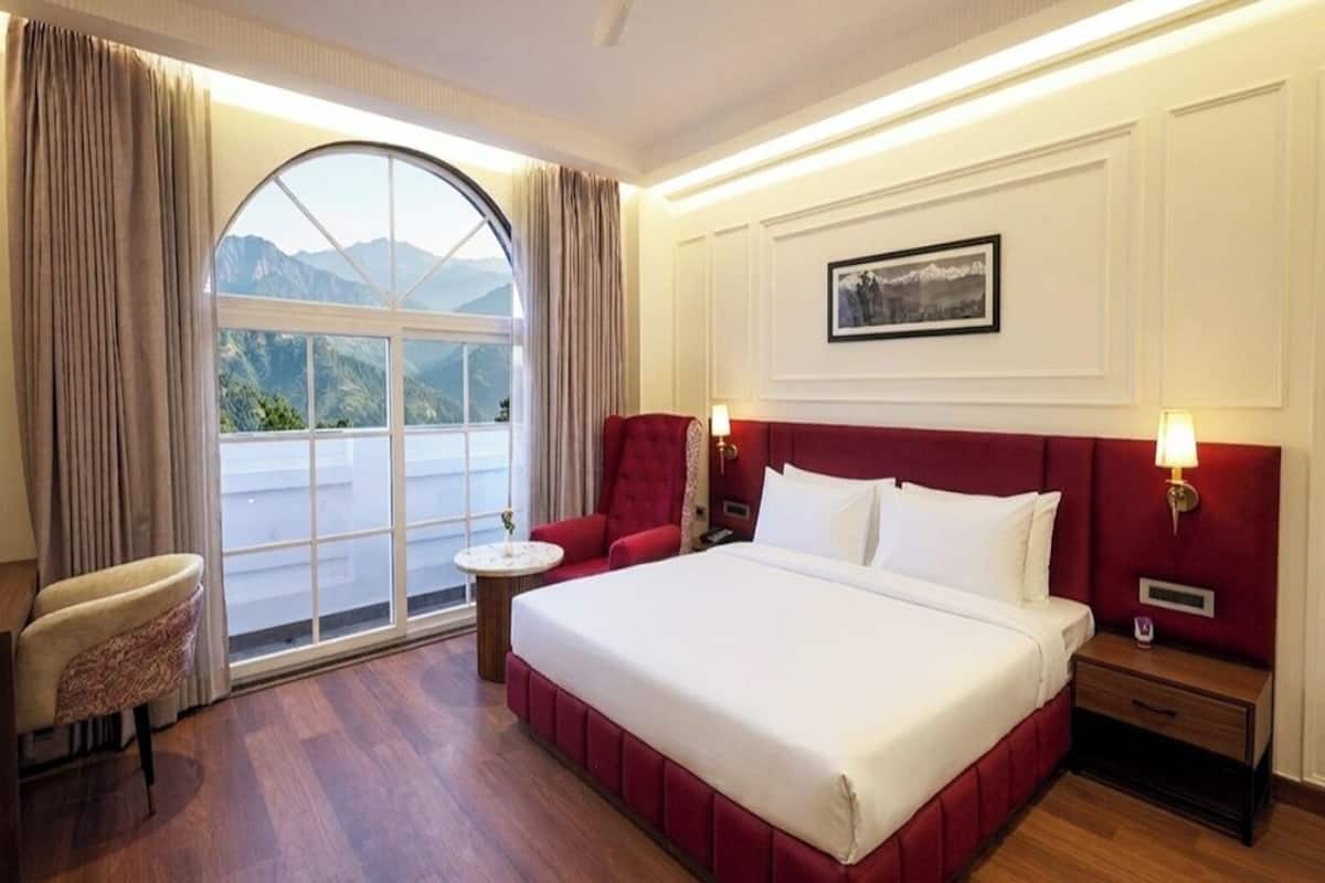 Habitación de hotel con una cama grande, cabecero rojo, mesita de noche, una pequeña mesa, una silla y un ventanal con vistas a las montañas.