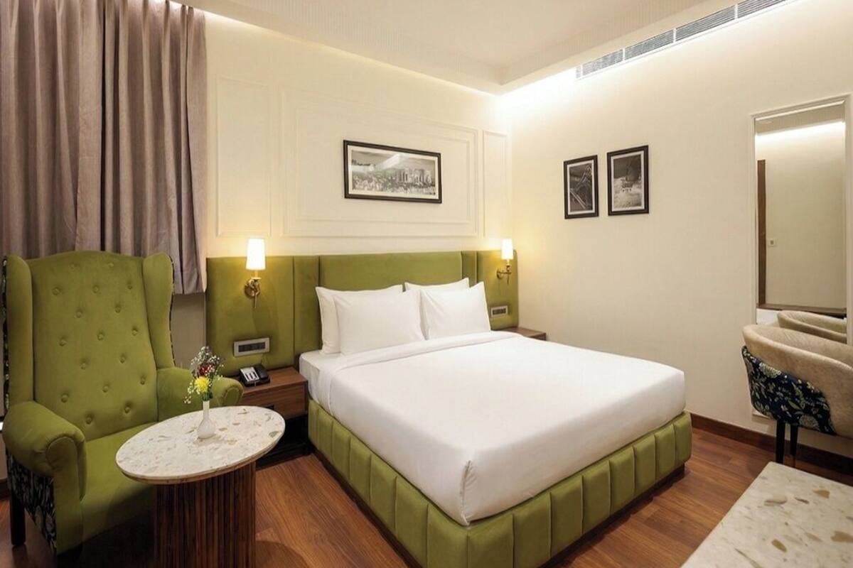 Habitación de hotel con un sillón tapizado en verde, una mesita redonda con un florero, una cama grande con sábanas blancas, dos marcos de cuadros en la pared y un espejo a la derecha.