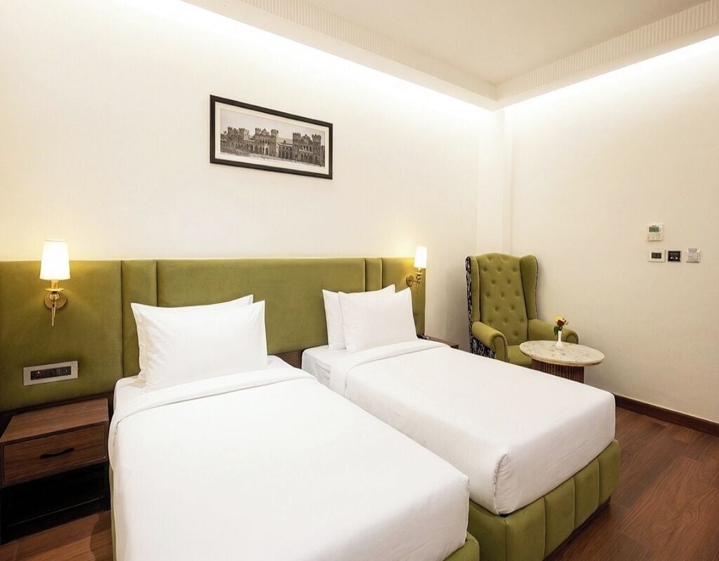 Habitación de hotel con dos camas, un cabecero tapizado en verde, un cuadro en la pared, un sillón verde, una mesita redonda y una mesita de noche con una lámpara.