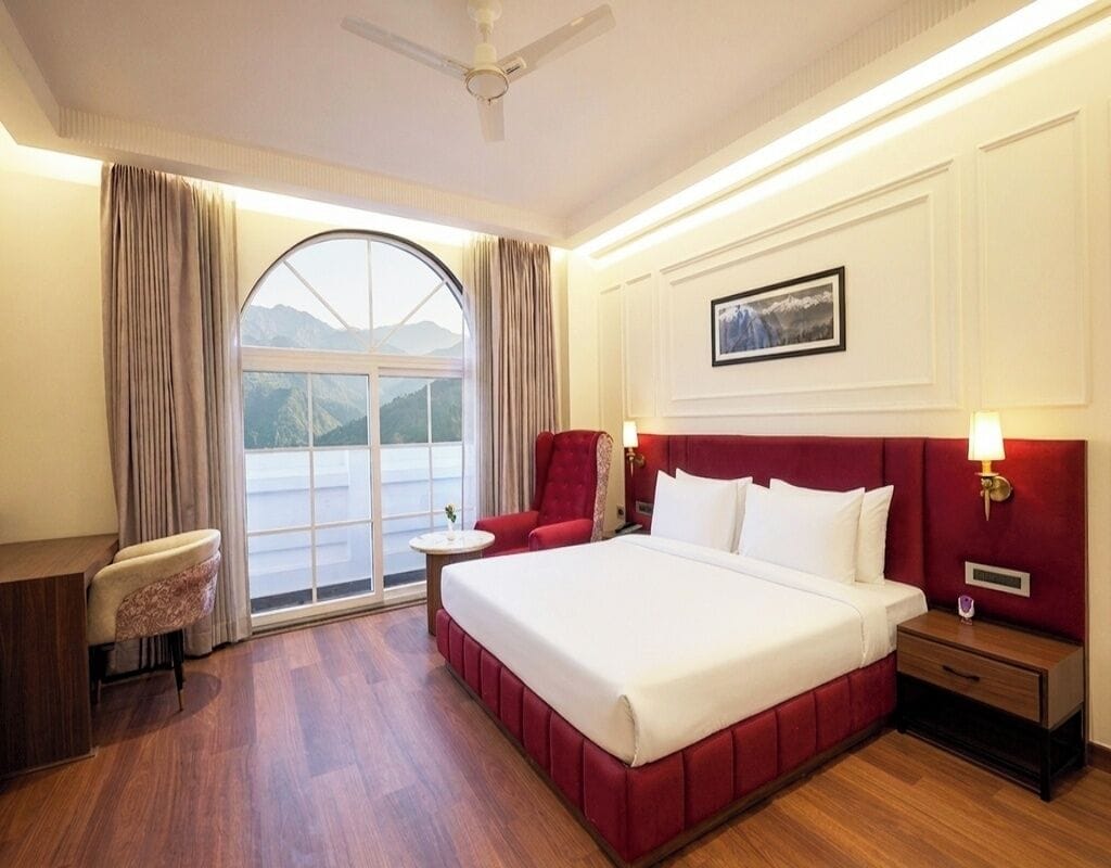 Habitación de hotel con cama, escritorio, silla, mesita de noche y ventana con vistas a las montañas.