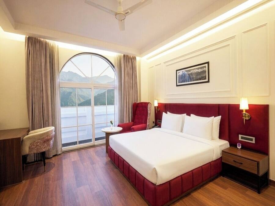 Habitación de hotel con cama, escritorio, silla, mesita de noche y ventana con vistas a las montañas.