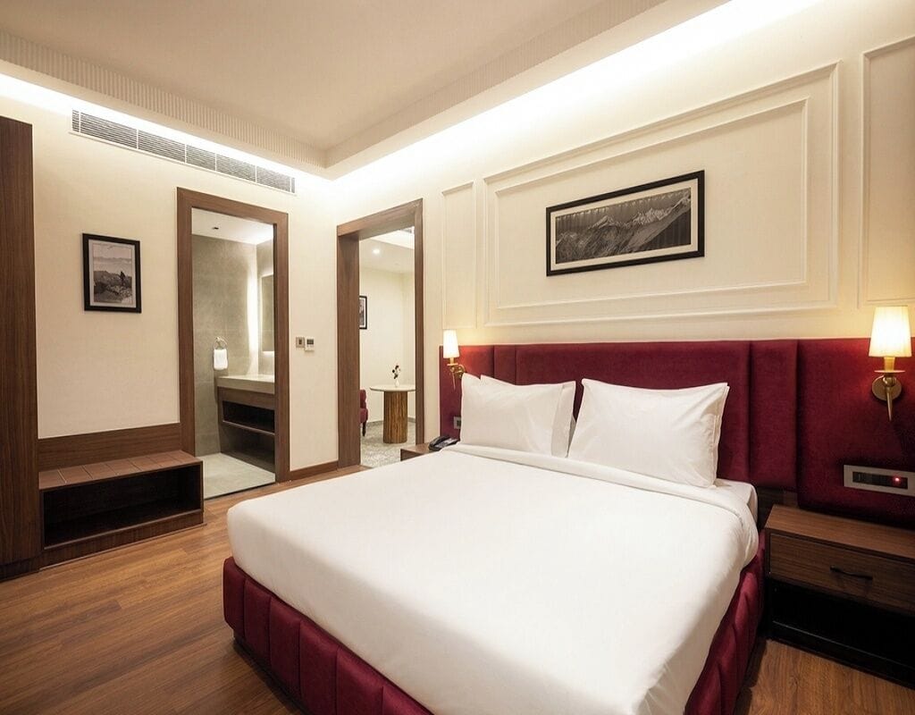 Habitación de hotel con una cama grande, un espejo, una mesita de noche con lámpara y un cuadro enmarcado en la pared.