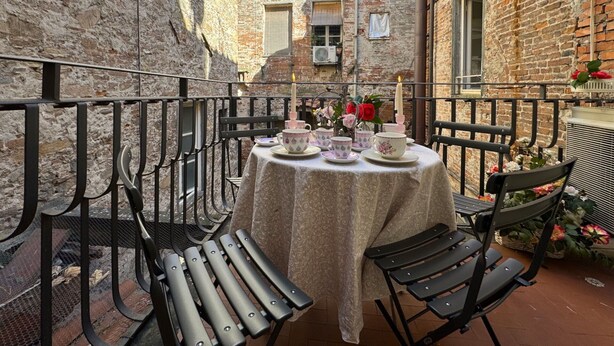 Une table dressée avec des tasses et des soucoupes à thé, une bougie et des fleurs, dans une cour extérieure aux murs de pierre et aux chaises en métal.