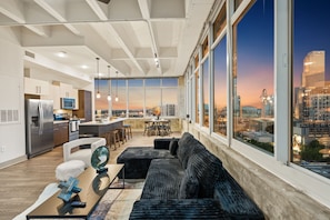 Eine moderne Küche mit Edelstahlgeräten, ein Essbereich mit einer schwarzen Couch und einem Tisch sowie große Fenster mit Blick auf die Stadt.