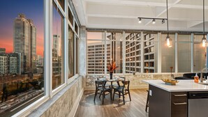 A modern kitchen with a large island, a dining area with a table and chairs, and a view of a cityscape through large windows. - Dual King Suites | Skyline Views | Downtown ATL (Atlanta)